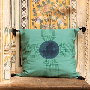 K&ouml;nnte beinhalten: Ein quadratisches, teal-farbenes Zierkissen mit einem dunkel-teal-farbenen kreisf&ouml;rmigen Design in der Mitte und halbkreisf&ouml;rmigen Mustern. Das Kissen hat schwarze Quasten an jeder Ecke. Das Kissen liegt auf einer Holzoberfl&auml;che.