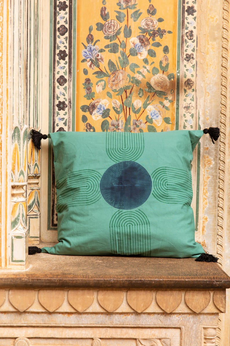 K&ouml;nnte beinhalten: Ein quadratisches, teal-farbenes Zierkissen mit einem dunkel-teal-farbenen kreisf&ouml;rmigen Design in der Mitte und halbkreisf&ouml;rmigen Mustern. Das Kissen hat schwarze Quasten an jeder Ecke. Das Kissen liegt auf einer Holzoberfl&auml;che.