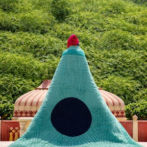 K&ouml;nnte beinhalten: Ein gro&szlig;es, t&uuml;rkisfarbenes, dreieckiges Textil mit einem schwarzen kreisf&ouml;rmigen Design. Das Textil ist &uuml;ber einer Struktur drapiert, mit einer roten und rosa Kopfbedeckung oben. Der Hintergrund zeigt &uuml;ppiges gr&uuml;nes Laub und architektonische Elemente.