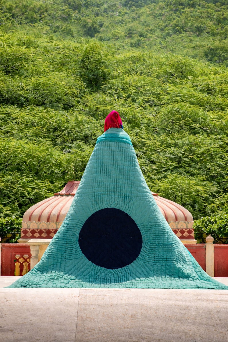 K&ouml;nnte beinhalten: Ein gro&szlig;es, t&uuml;rkisfarbenes, dreieckiges Textil mit einem schwarzen kreisf&ouml;rmigen Design. Das Textil ist &uuml;ber einer Struktur drapiert, mit einer roten und rosa Kopfbedeckung oben. Der Hintergrund zeigt &uuml;ppiges gr&uuml;nes Laub und architektonische Elemente.