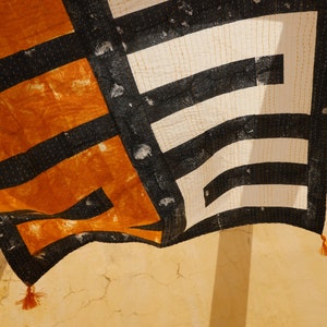 May include: A black and white patterned fabric with orange accents hangs from a wall. The fabric has a geometric design and is sewn with a running stitch. The fabric has tassels on the bottom corners.