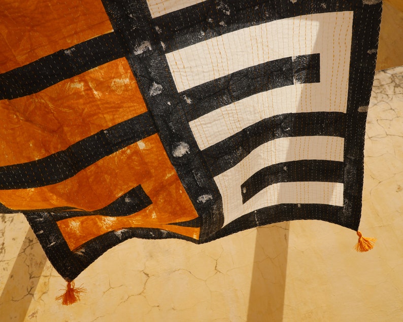 May include: A black and white patterned fabric with orange accents hangs from a wall. The fabric has a geometric design and is sewn with a running stitch. The fabric has tassels on the bottom corners.