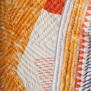 May include: Close-up of a quilted textile with a patchwork design. The quilt features sections of orange, white, and red fabric. The orange sections have a tie-dye effect, while the white sections have a textured, quilted pattern.