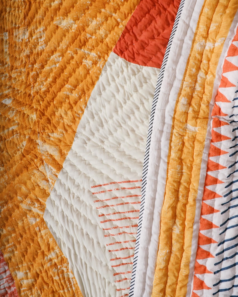 May include: Close-up of a quilted textile with a patchwork design. The quilt features sections of orange, white, and red fabric. The orange sections have a tie-dye effect, while the white sections have a textured, quilted pattern.