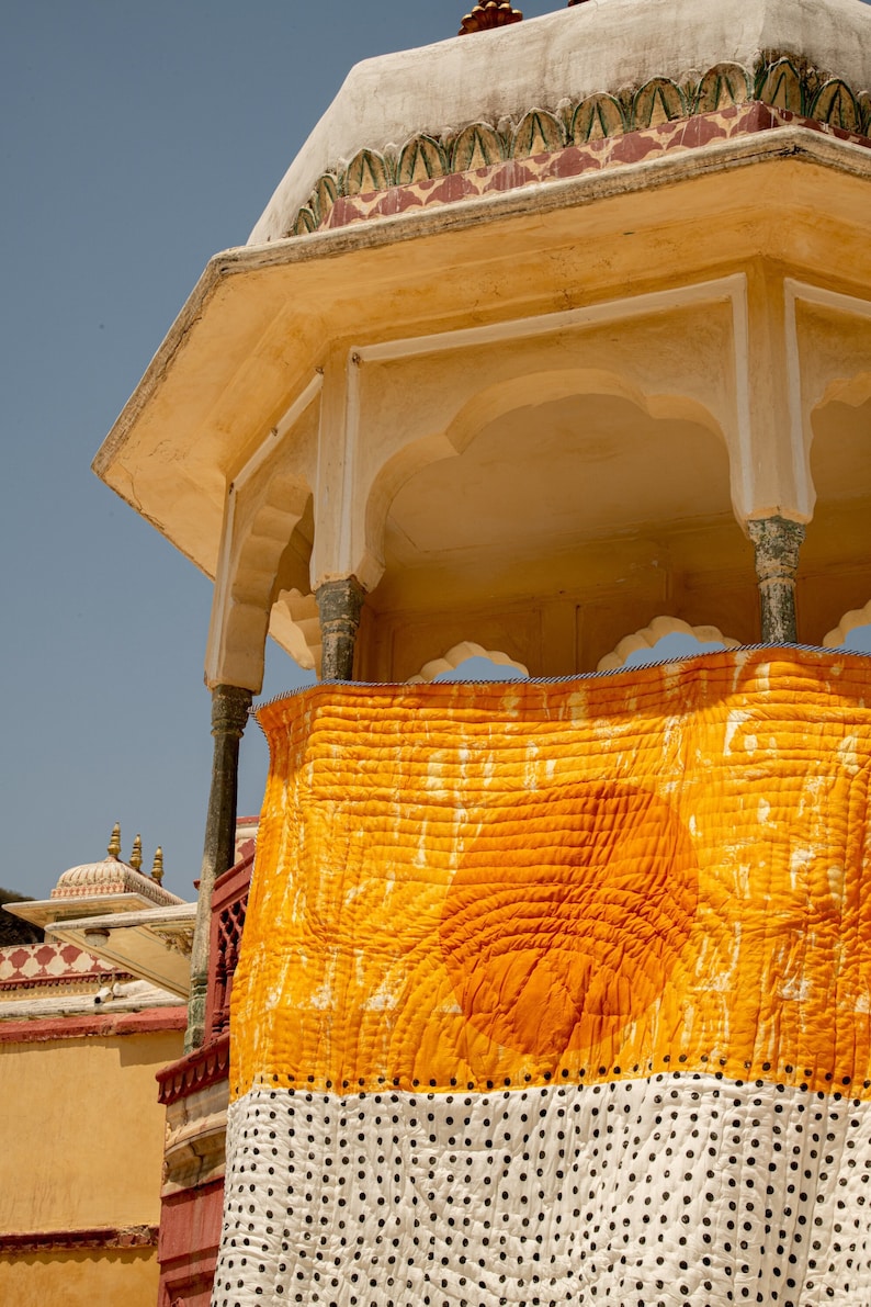 May include: A yellow and white patterned quilt hangs from a yellow building with a white trim. The quilt has a large yellow section with a white pattern and a white section with black polka dots.
