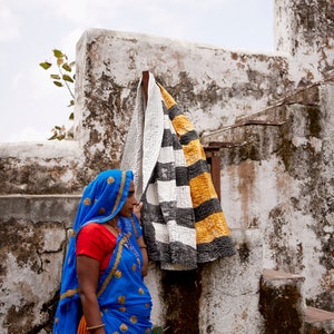 May include: A woman wearing a blue and orange sari stands in front of a whitewashed wall. A black and white striped blanket with yellow accents hangs on the wall behind her.