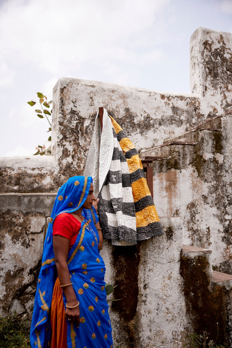 May include: A woman wearing a blue and orange sari stands in front of a whitewashed wall. A black and white striped blanket with yellow accents hangs on the wall behind her.