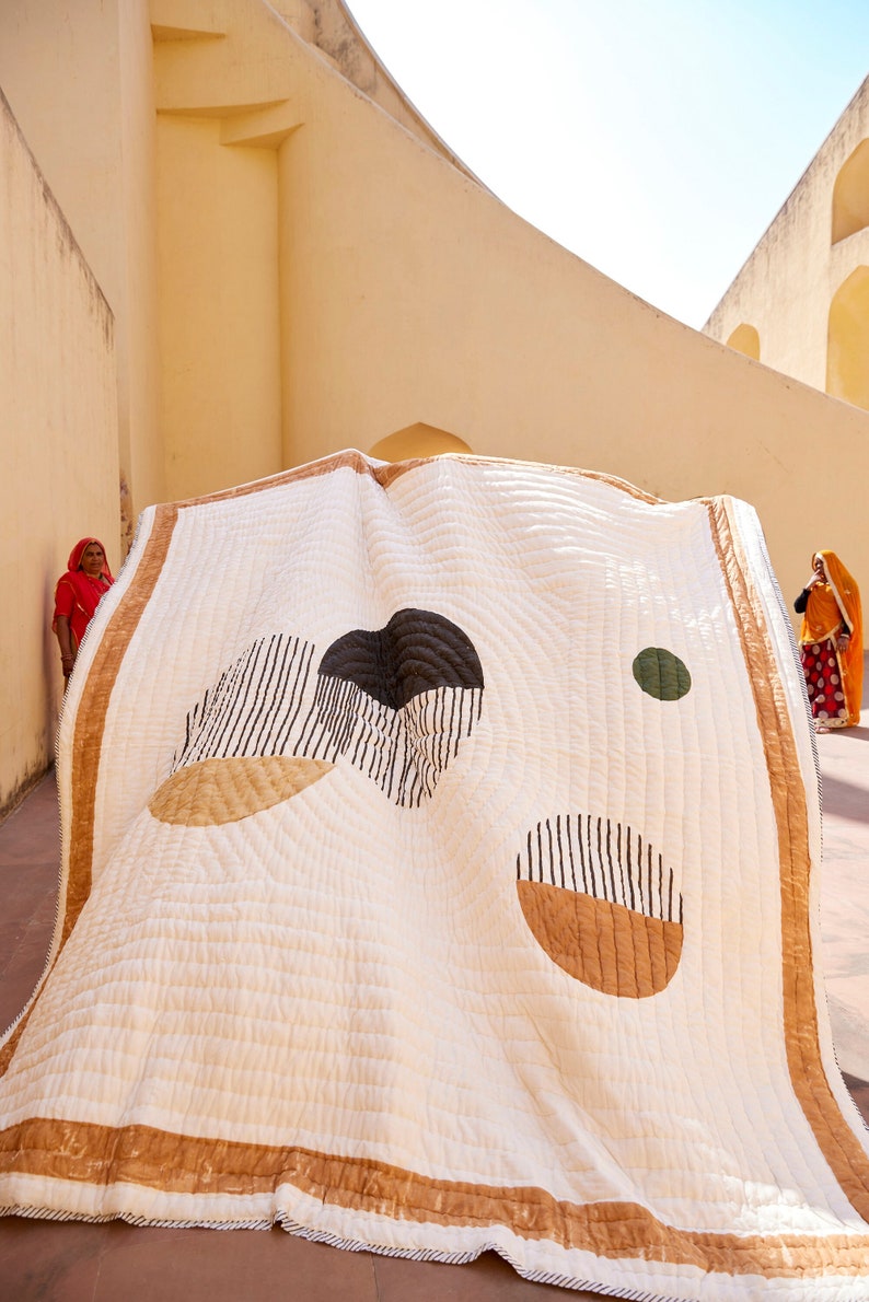 May include: A white cotton quilt with a geometric design featuring black, brown, and green shapes. The quilt is folded and displayed in front of a yellow wall.