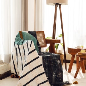 Op de afbeelding: Een wit en zwart gestreepte deken met oranje stiksels en kwastjes, gedrapeerd over een houten stoel met een gevlochten rieten zitting. Een houten staande lamp met een beige lampenkap staat achter de stoel.