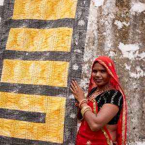 May include: A woman wearing a red sari with gold embroidery and a black blouse stands in front of a yellow and black patterned fabric. She is smiling and looking at the camera.