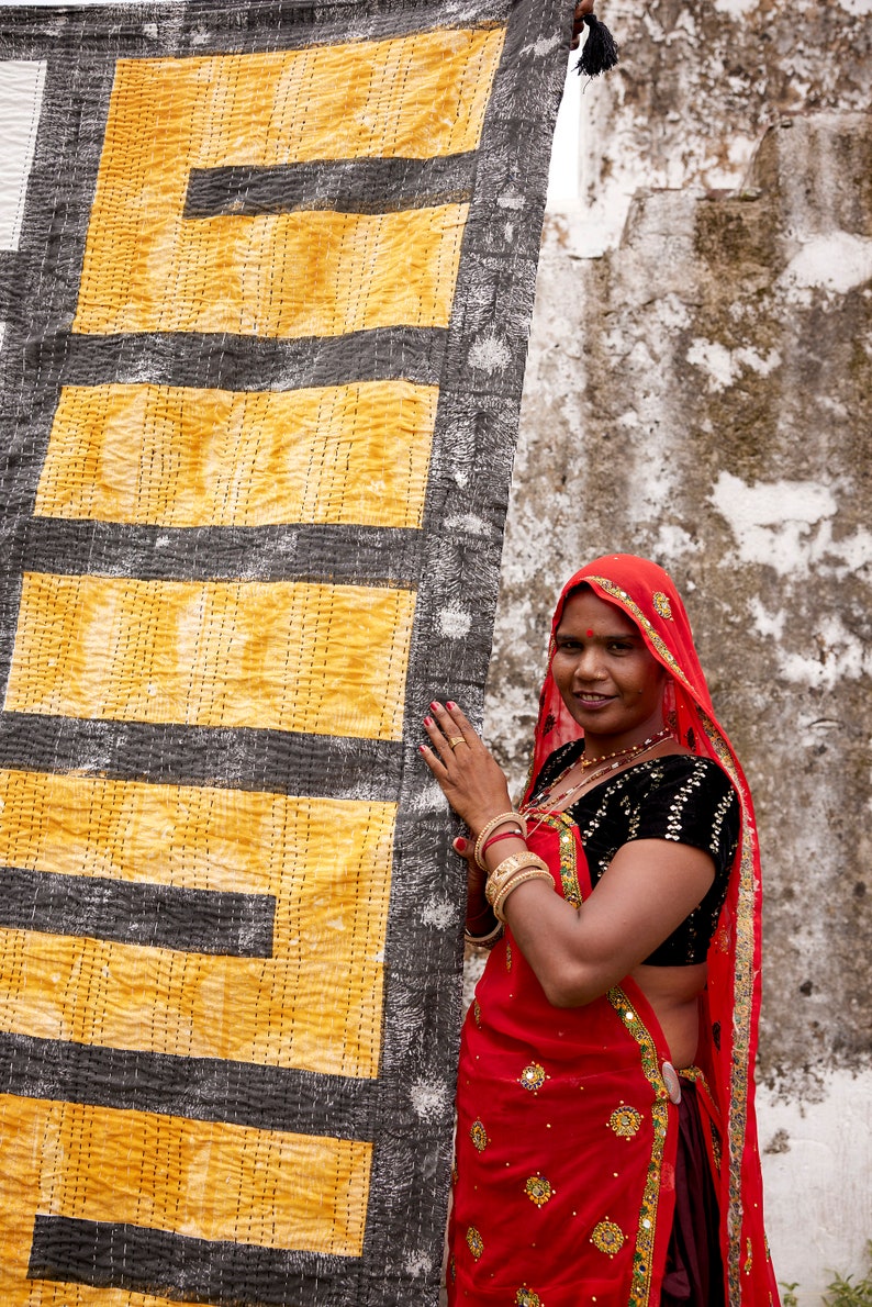 May include: A woman wearing a red sari with gold embroidery and a black blouse stands in front of a yellow and black patterned fabric. She is smiling and looking at the camera.