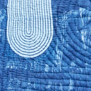 K&ouml;nnte beinhalten: Nahaufnahme eines blau-wei&szlig;en Quilts mit einzigartigem Design. Der Quilt zeigt eine gro&szlig;e, gebogene wei&szlig;e Form mit vertikalen blauen Linien, vor einem strukturierten, indigogef&auml;rbten Hintergrund. Das Steppmuster verleiht Tiefe.