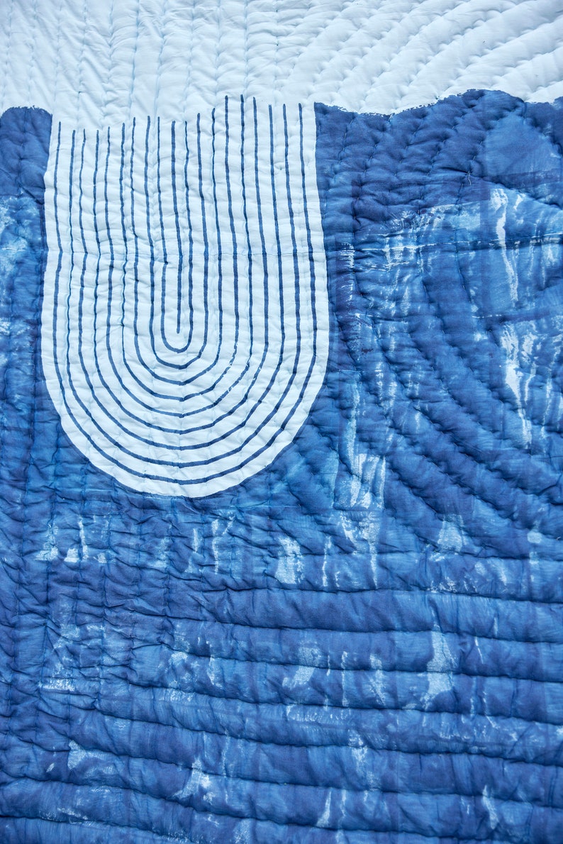 K&ouml;nnte beinhalten: Nahaufnahme eines blau-wei&szlig;en Quilts mit einzigartigem Design. Der Quilt zeigt eine gro&szlig;e, gebogene wei&szlig;e Form mit vertikalen blauen Linien, vor einem strukturierten, indigogef&auml;rbten Hintergrund. Das Steppmuster verleiht Tiefe.