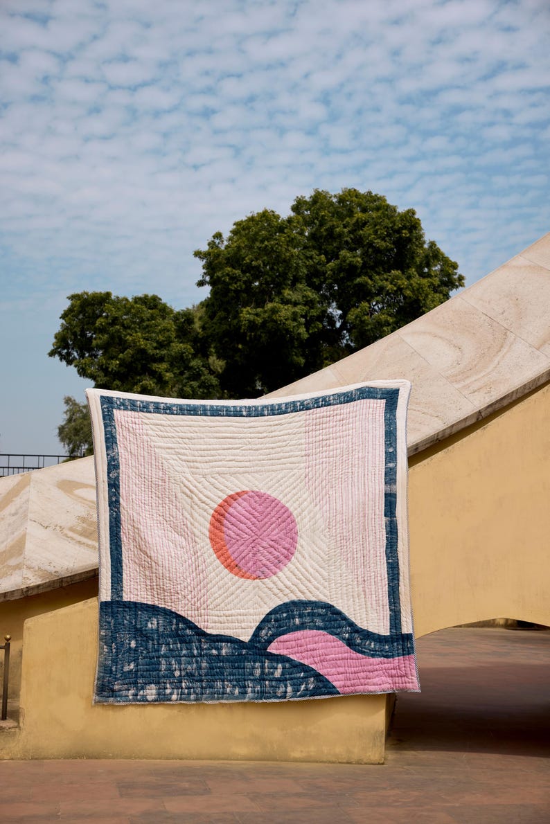 May include: A quilted textile with a geometric design. The quilt features a pink and orange sun, blue and pink landscape elements, and a blue border on a cream background. The quilt is hanging outdoors against a blue sky and trees.
