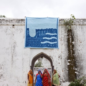 K&ouml;nnte beinhalten: Ein blau-wei&szlig; gestepptes Wandbehang mit Wellenmuster wird an einer wei&szlig;en Wand gezeigt. Vier Frauen in farbenfroher traditioneller Kleidung stehen vor einem gew&ouml;lbten Tor. Die Frauen tragen Saris in Orange-, Blau-, Rot- und Gr&uuml;nt&ouml;nen.