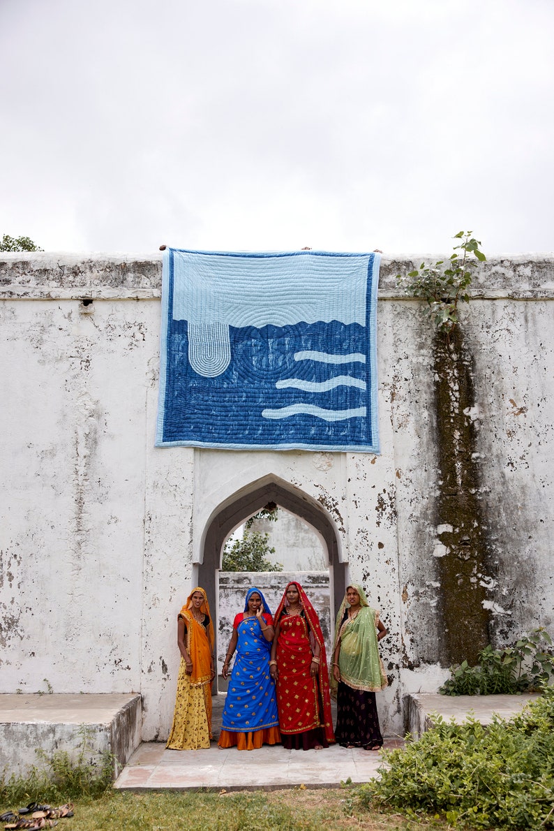 K&ouml;nnte beinhalten: Ein blau-wei&szlig; gestepptes Wandbehang mit Wellenmuster wird an einer wei&szlig;en Wand gezeigt. Vier Frauen in farbenfroher traditioneller Kleidung stehen vor einem gew&ouml;lbten Tor. Die Frauen tragen Saris in Orange-, Blau-, Rot- und Gr&uuml;nt&ouml;nen.