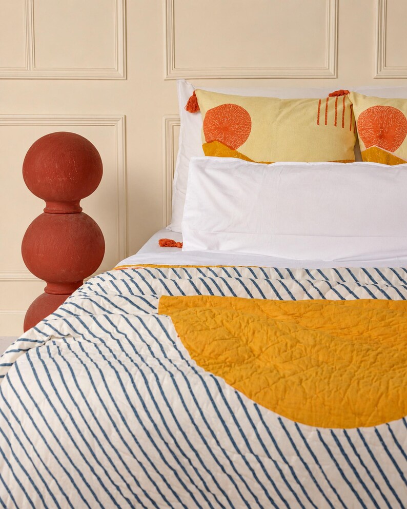 May include: A bed with white sheets and a striped comforter featuring a yellow half-circle design. Two yellow pillows with orange sunburst patterns and tassels are visible. A tall, red decorative object stands to the left.