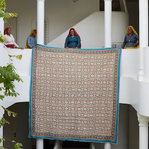 Puede incluir: Cuatro mujeres se paran en un balc&oacute;n blanco sosteniendo una gran colcha colorida con un patr&oacute;n geom&eacute;trico. La colcha est&aacute; hecha de una variedad de telas y tiene un borde azul. Las mujeres visten ropa tradicional india.