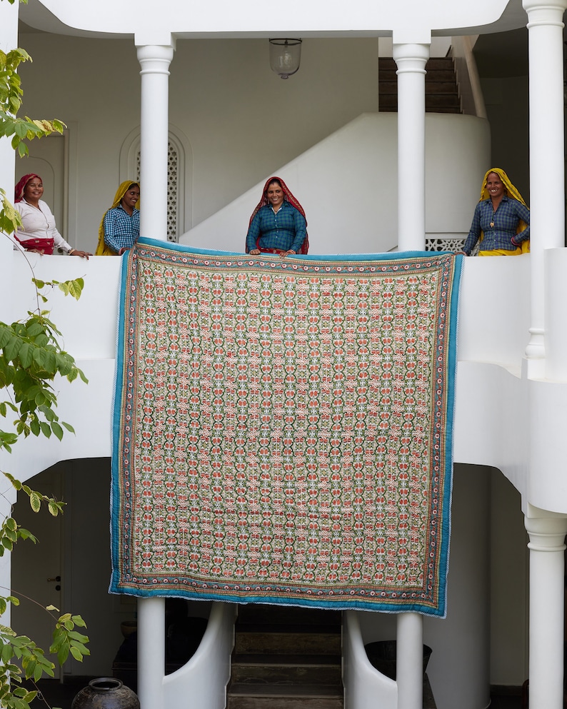 Puede incluir: Cuatro mujeres se paran en un balc&oacute;n blanco sosteniendo una gran colcha colorida con un patr&oacute;n geom&eacute;trico. La colcha est&aacute; hecha de una variedad de telas y tiene un borde azul. Las mujeres visten ropa tradicional india.