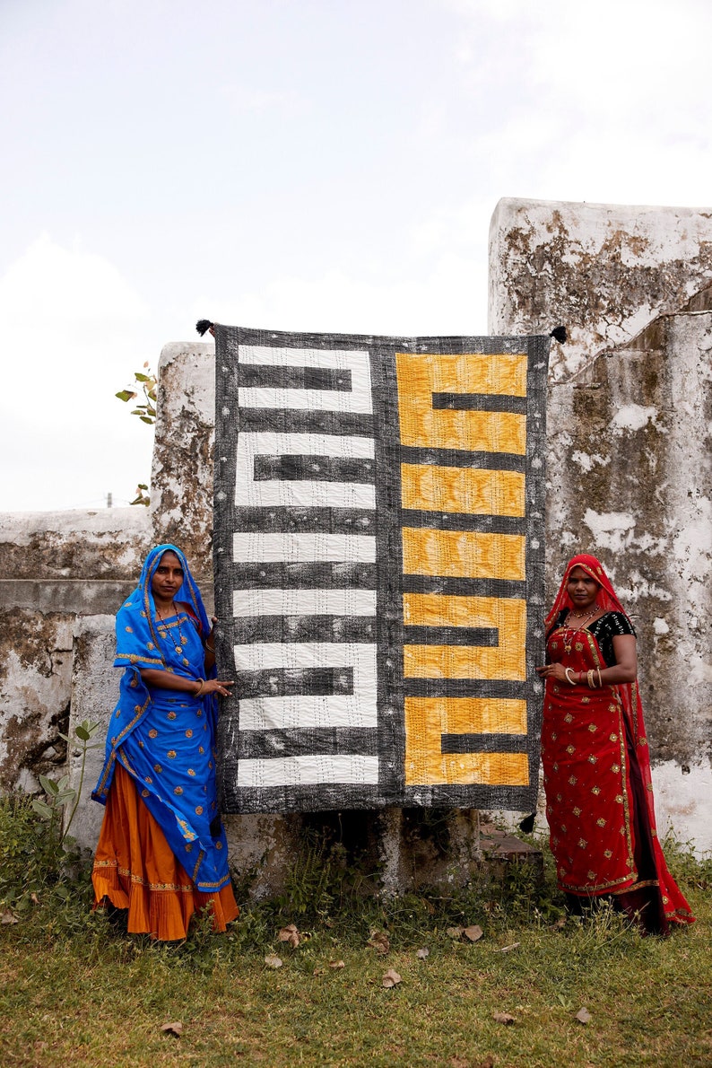 May include: Two people stand in front of a large, black and white patterned fabric with a yellow section. The fabric has a geometric design. The person on the left is wearing a blue and orange sari. The person on the right is wearing a red sari.