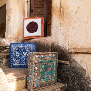 May include: Three colorful patterned floor cushions sit on a set of stone steps in front of a building. The cushions are square and have a variety of floral and geometric designs. The top cushion has a large brown circle in the center.