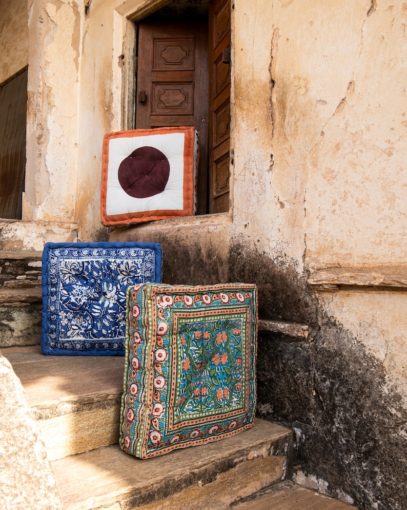 May include: Three colorful patterned floor cushions sit on a set of stone steps in front of a building. The cushions are square and have a variety of floral and geometric designs. The top cushion has a large brown circle in the center.