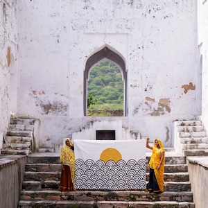 Peut inclure: Un textile rectangulaire pr&eacute;sentant une impression graphique d'un soleil jaune se levant sur un motif r&eacute;p&eacute;titif de vagues oc&eacute;aniques stylis&eacute;es en noir et blanc. Le tissu est tenu horizontalement devant des marches en pierre blanche.