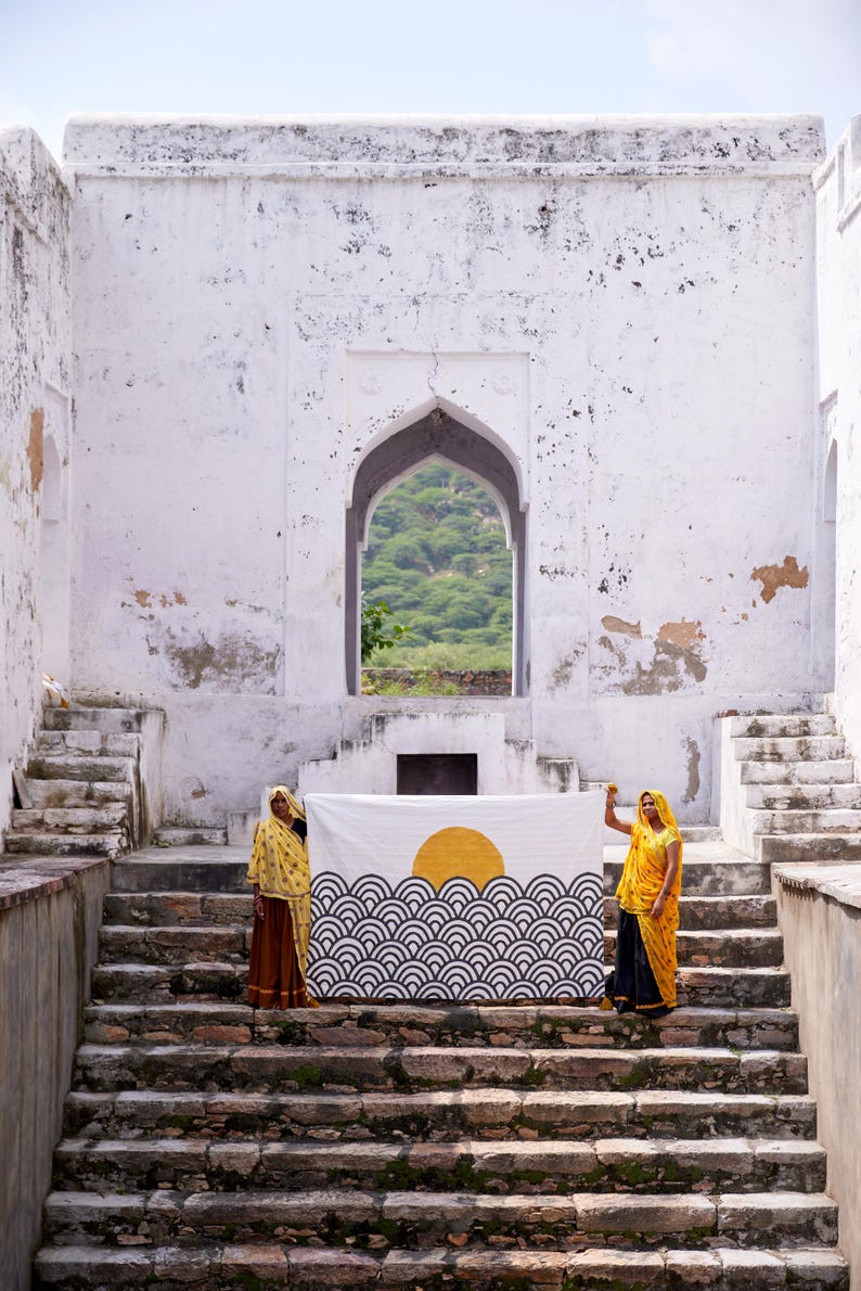 Peut inclure: Un textile rectangulaire pr&eacute;sentant une impression graphique d'un soleil jaune se levant sur un motif r&eacute;p&eacute;titif de vagues oc&eacute;aniques stylis&eacute;es en noir et blanc. Le tissu est tenu horizontalement devant des marches en pierre blanche.