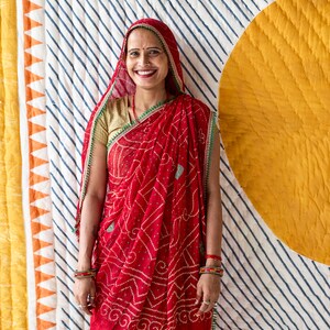 May include: A woman wearing a red sari with a white and red patterned design. The sari has a green border and is paired with a gold blouse and a red head covering. The background features a striped pattern and a yellow circle.