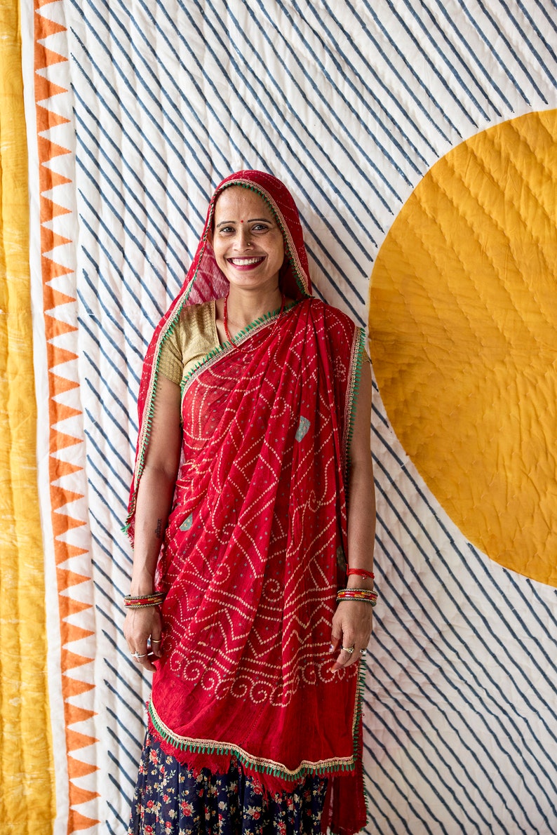 May include: A woman wearing a red sari with a white and red patterned design. The sari has a green border and is paired with a gold blouse and a red head covering. The background features a striped pattern and a yellow circle.