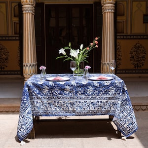 Puede incluir: Un mantel de mesa azul y blanco con un dise&ntilde;o floral est&aacute; preparado para una cena para dos. Dos cubiertos est&aacute;n dispuestos con platos blancos, servilletas rosas y vasos transparentes. Un ramo de flores blancas en un jarr&oacute;n transparente se encuentra en el centro de la mesa.