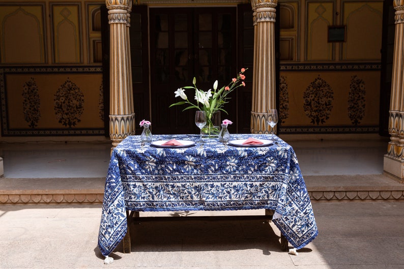 Puede incluir: Un mantel de mesa azul y blanco con un dise&ntilde;o floral est&aacute; preparado para una cena para dos. Dos cubiertos est&aacute;n dispuestos con platos blancos, servilletas rosas y vasos transparentes. Un ramo de flores blancas en un jarr&oacute;n transparente se encuentra en el centro de la mesa.