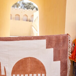 May include: A white rug with a brown geometric pattern. The rug has a fringe border and is hanging on a wall.