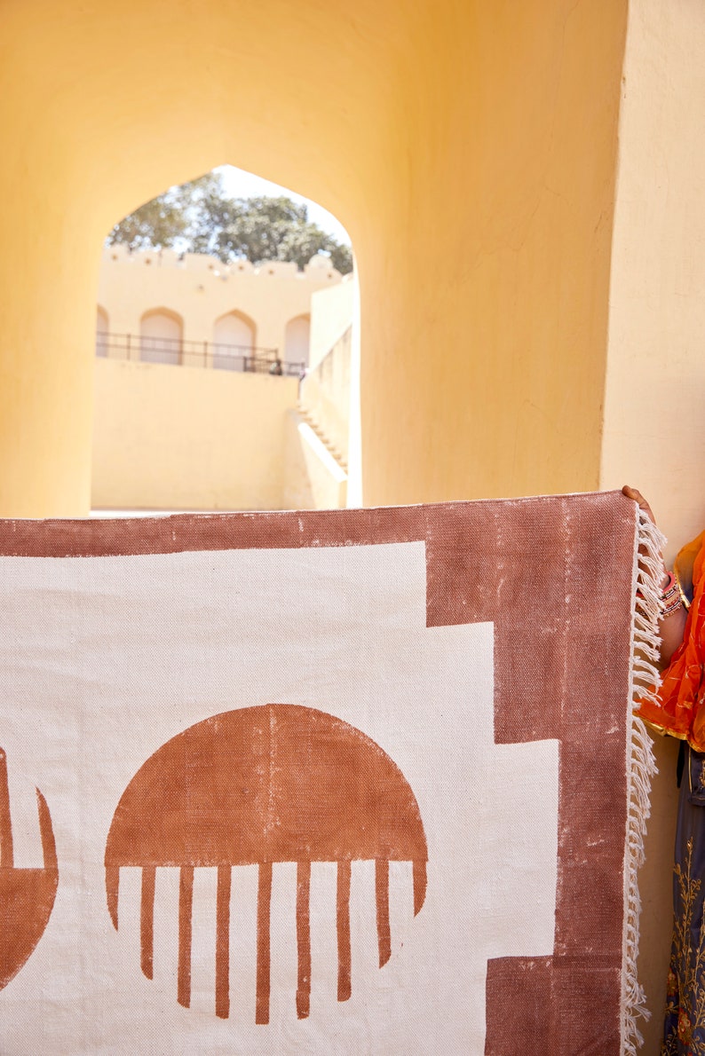 May include: A white rug with a brown geometric pattern. The rug has a fringe border and is hanging on a wall.