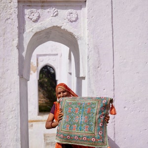 Pu&ograve; includere: Una donna con un foulard rosso e arancione sorride mentre tiene un cuscino con motivi colorati e frange. Il cuscino &egrave; davanti a un arco bianco.