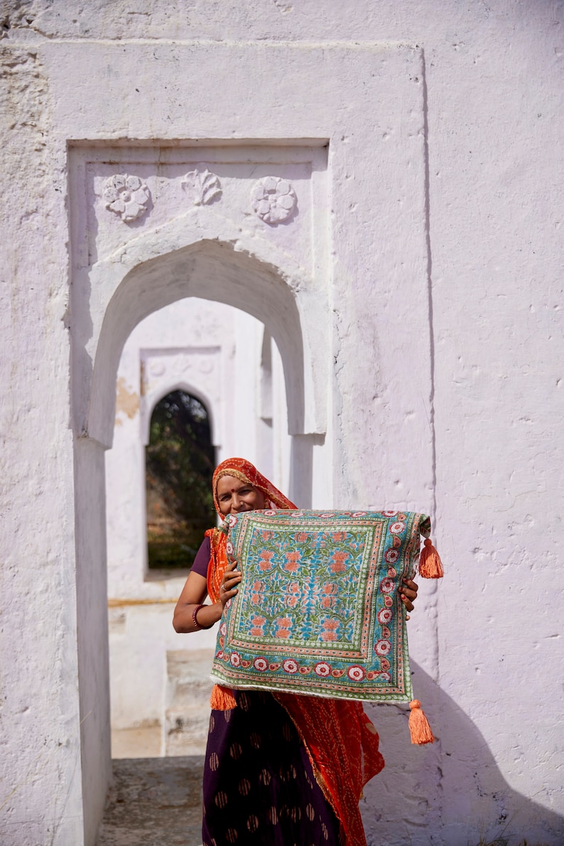 Pu&ograve; includere: Una donna con un foulard rosso e arancione sorride mentre tiene un cuscino con motivi colorati e frange. Il cuscino &egrave; davanti a un arco bianco.