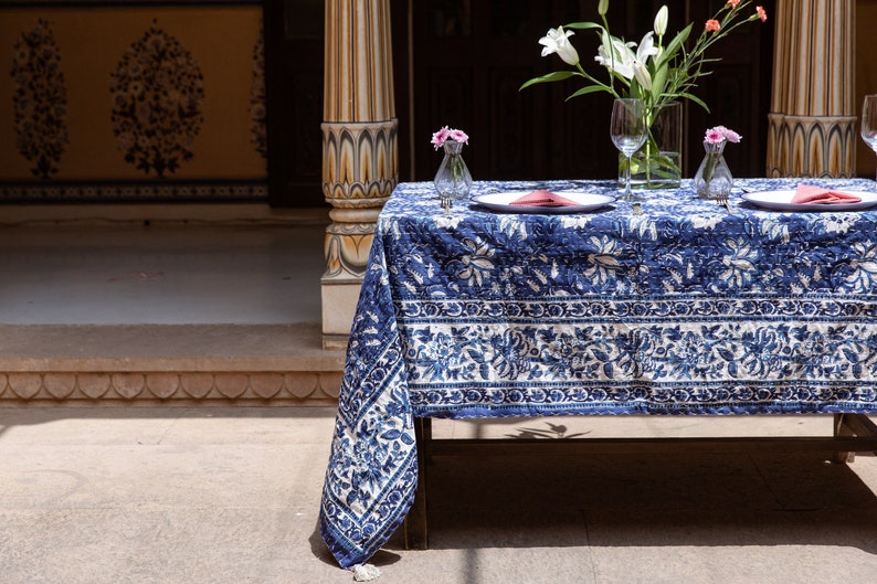 Puede incluir: Un mantel de mesa con estampado azul y blanco con un dise&ntilde;o floral, colocado sobre una mesa con dos cubiertos, cada uno con un plato blanco, una servilleta rosa y un vaso de agua. El mantel tiene un borde con flecos.