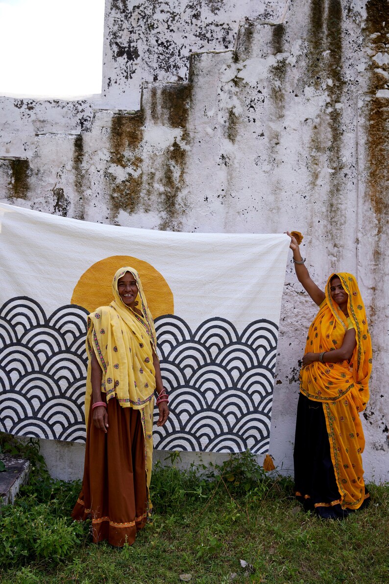 Peut inclure: Un textile rectangulaire blanc pr&eacute;sentant un grand motif de soleil jaune au-dessus de motifs de vagues en arc gris fonc&eacute; r&eacute;p&eacute;titifs. Le tissu pr&eacute;sente des coutures complexes sur toute sa surface, cr&eacute;ant une texture au design graphique &eacute;pur&eacute;.
