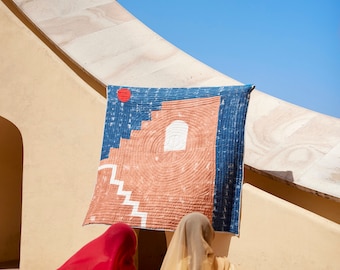 May include: A colorful textile art piece with a geometric design is displayed against a beige wall. The artwork features a blue and orange color scheme with a red circle and white stairs. Two women in traditional attire stand in front of the wall.