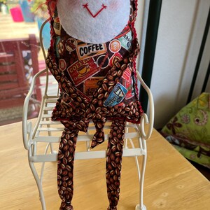 May include: A fabric doll with a white felt face, button eyes, and a red stitched mouth, wearing a brown coffee-themed patterned outfit with a coffee bean print. The doll is seated on a white metal chair with a slatted seat and curved backrest.
