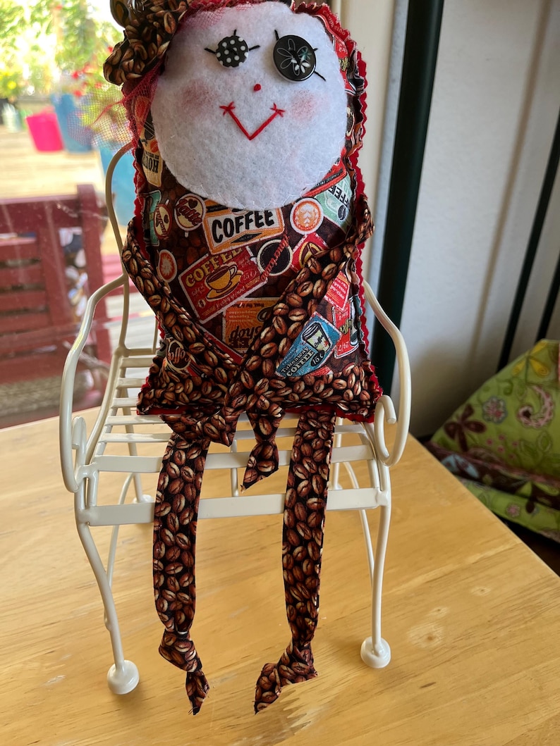 May include: A fabric doll with a white felt face, button eyes, and a red stitched mouth, wearing a brown coffee-themed patterned outfit with a coffee bean print. The doll is seated on a white metal chair with a slatted seat and curved backrest.