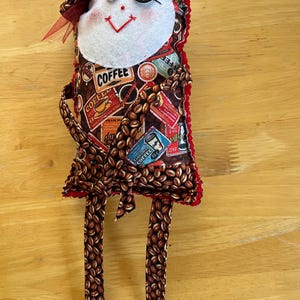 May include: A handmade fabric doll featuring a white felt face with button eyes, a red stitched mouth, and a red tulle bow. The body is covered in a brown coffee bean pattern with various coffee-themed labels and text, including the word COFFEE.
