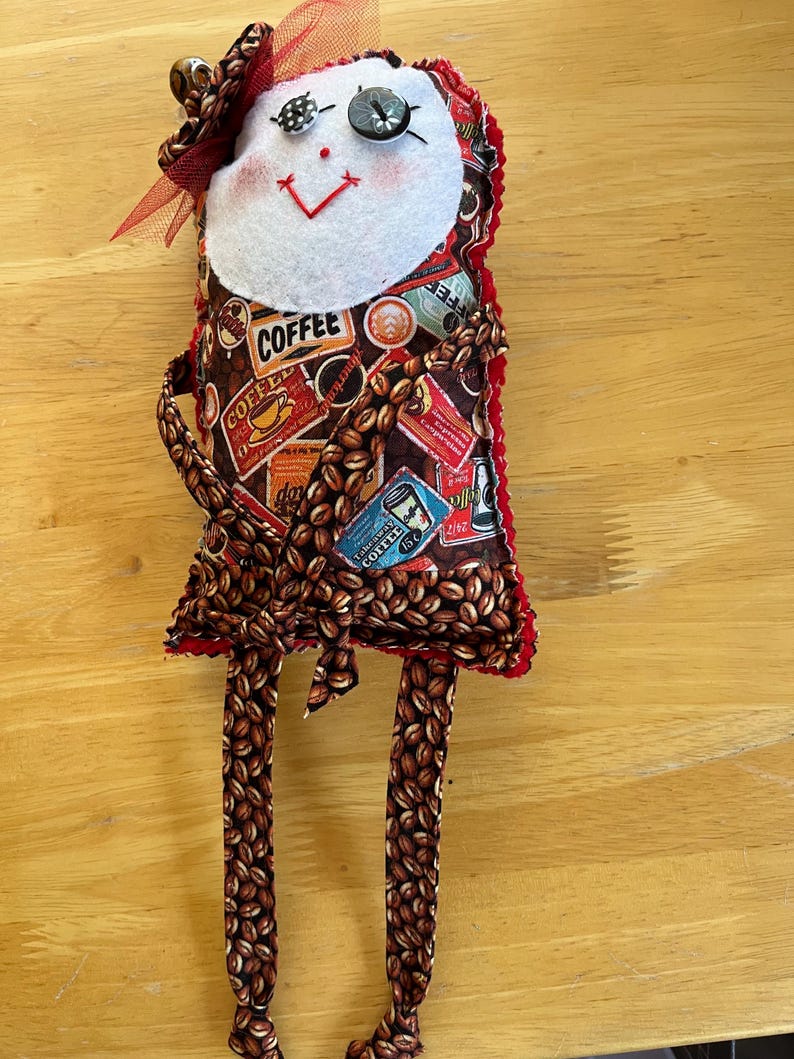 May include: A handmade fabric doll featuring a white felt face with button eyes, a red stitched mouth, and a red tulle bow. The body is covered in a brown coffee bean pattern with various coffee-themed labels and text, including the word COFFEE.