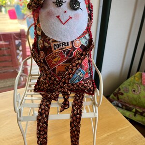 May include: A handmade fabric doll with a white felt face, button eyes, and a red stitched mouth. The body features a brown coffee bean pattern with coffee-themed text and graphics. The doll wears a matching fabric bow and sits on a white metal chair.