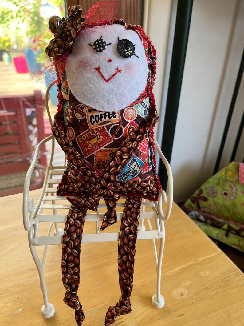 May include: A handmade fabric doll with a white felt face, button eyes, and a red stitched mouth. The body features a brown coffee bean pattern with coffee-themed text and graphics. The doll wears a matching fabric bow and sits on a white metal chair.