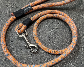 May include: A dog lead made of thick, braided rope in a vibrant orange colour with small blue flecks throughout. The lead features a silver-toned metal swivel snap hook and a black fabric tag with the white text ObieDog attached to the rope.