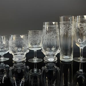 May include: A collection of six clear glass drinking glasses with etched floral designs. The glasses vary in size and shape, including stemmed wine glasses and tall tumblers. The glasses are arranged on a reflective black surface.