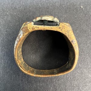 Puede incluir: Un anillo de lat&oacute;n antiguo y desgastado con un dise&ntilde;o floral en la parte superior. El anillo tiene un aspecto deslustrado y envejecido, con desgaste y decoloraci&oacute;n visibles. La banda es de forma ovalada y el elemento floral est&aacute; ligeramente elevado.