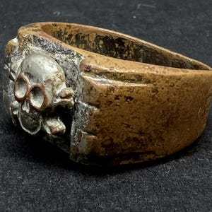 Puede incluir: Un anillo vintage con dise&ntilde;o de calavera. El anillo presenta una calavera con cuencas de ojos y dientes visibles, engastada sobre una banda met&aacute;lica desgastada. El anillo tiene un aspecto deslustrado y envejecido, con una mezcla de tonos plateados y bronce.