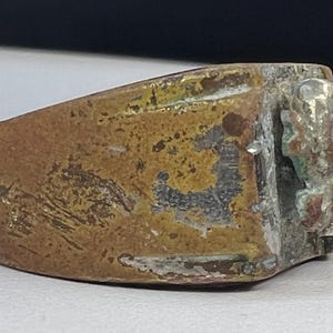 Puede incluir: Un anillo desgastado de color bronce con un dise&ntilde;o detallado, posiblemente antiguo, en la cara. El anillo muestra signos de edad y desgaste, con una apariencia deslustrada y textura visible. El dise&ntilde;o parece ser un relieve.